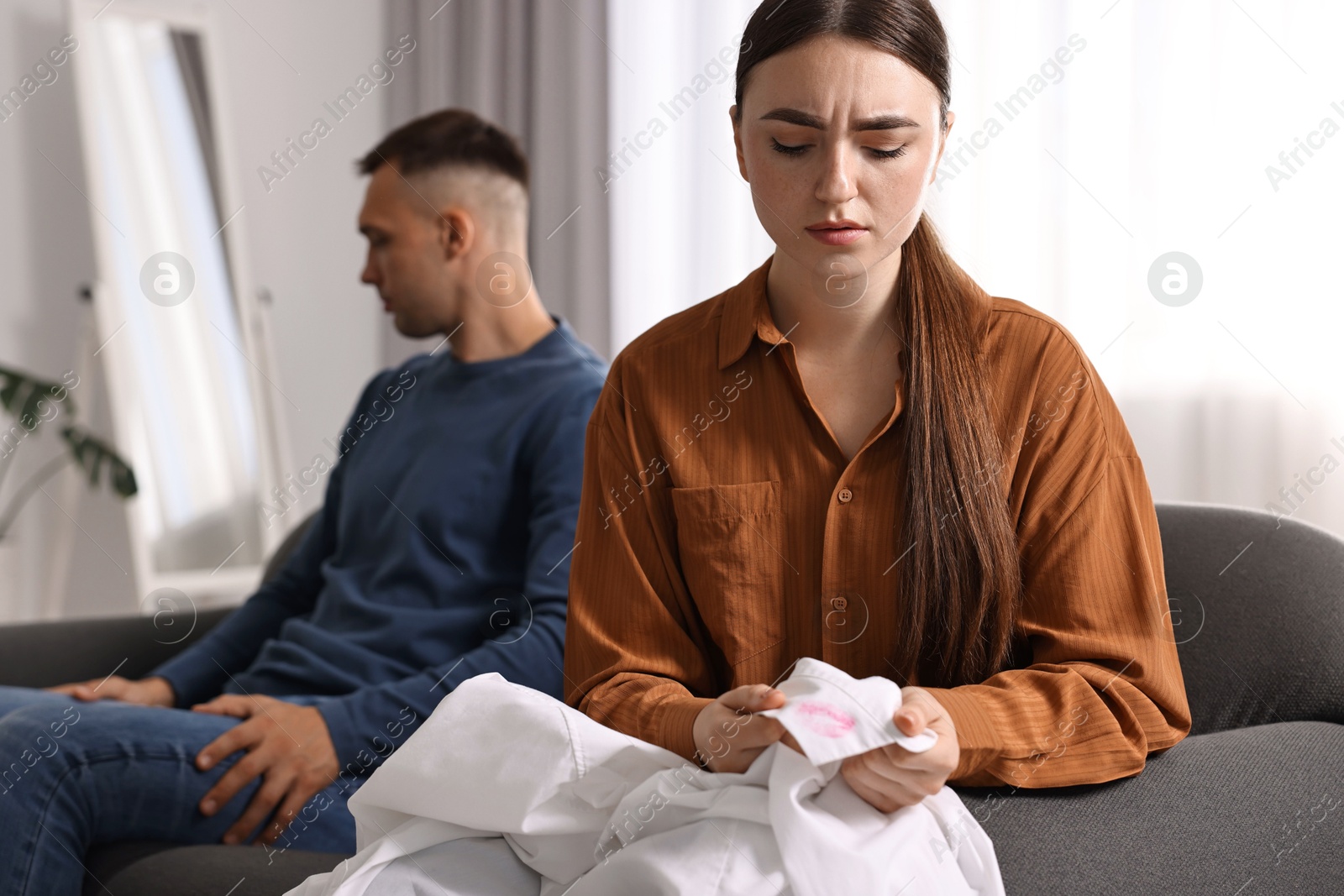 Photo of Frustrated wife holding shirt with lipstick marks as proof of her unfaithful husband at home, selective focus