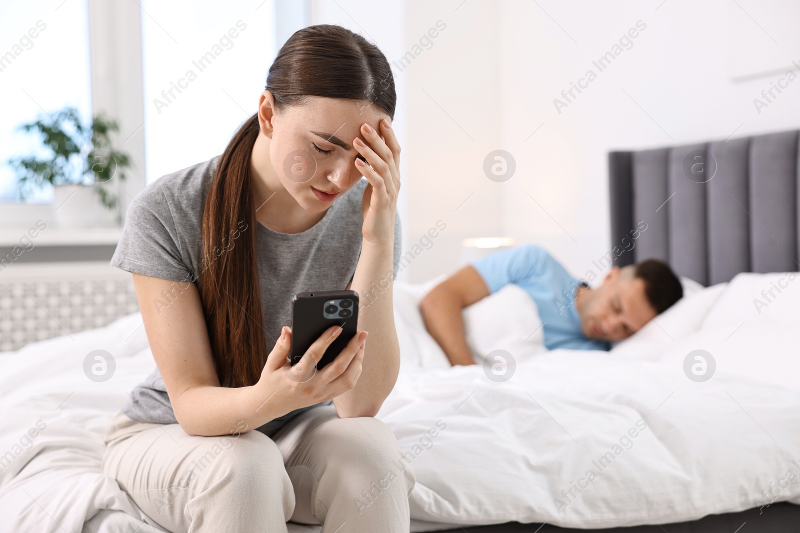 Photo of Frustrated woman with husband's smartphone while he sleeping in bedroom, selective focus