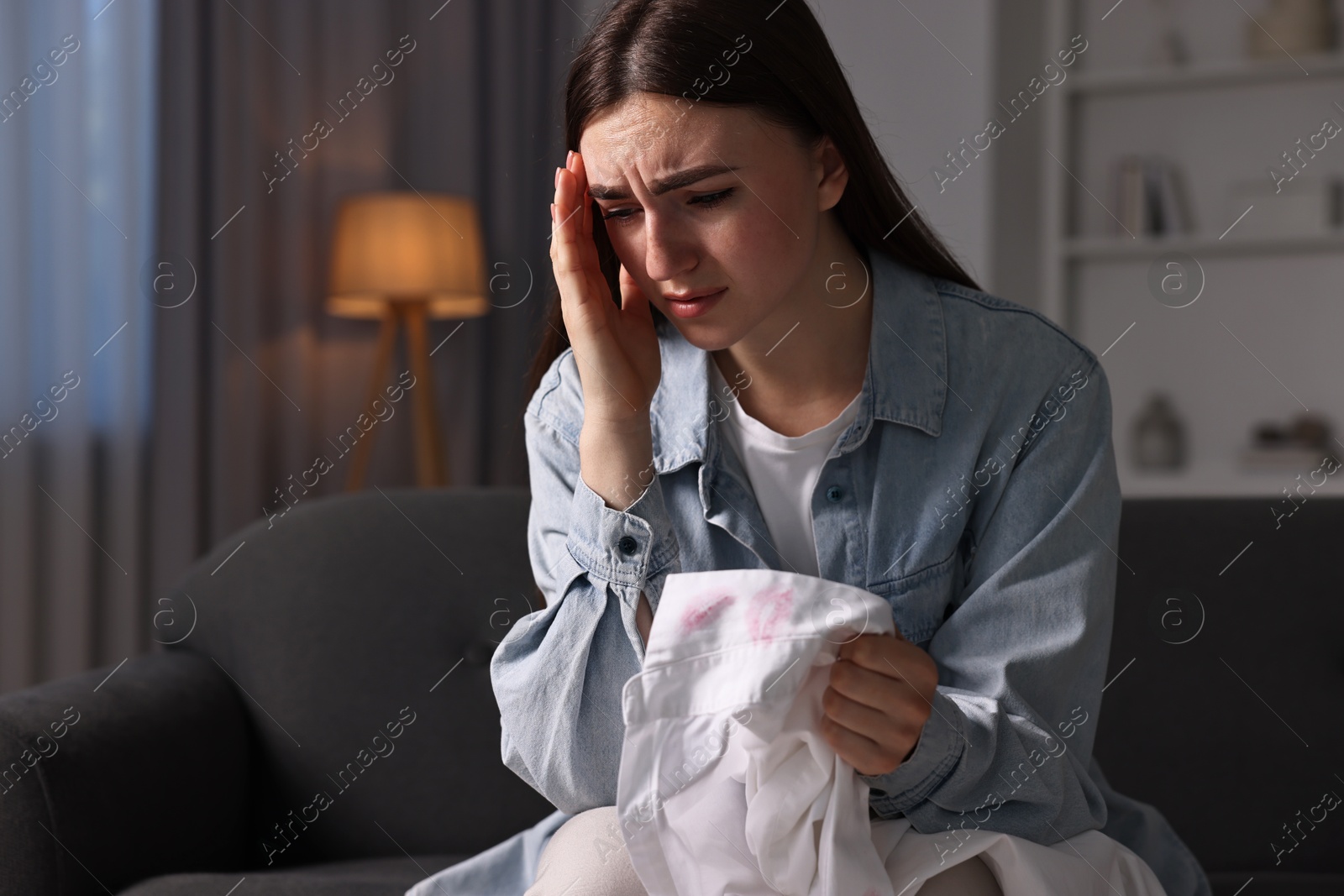 Photo of Frustrated wife holding shirt with lipstick marks as proof of her unfaithful husband at home