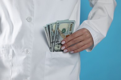 Photo of Corruption concept. Doctor putting dollar banknotes into her pocket on light blue background, closeup