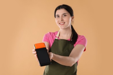 Happy young woman in apron with payment terminal on beige background