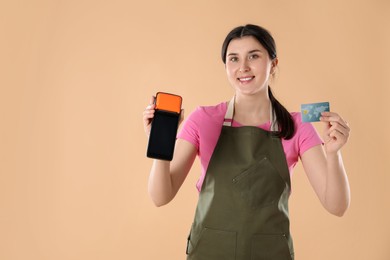 Photo of Happy young woman in apron with payment terminal and debit card on beige background, space for text