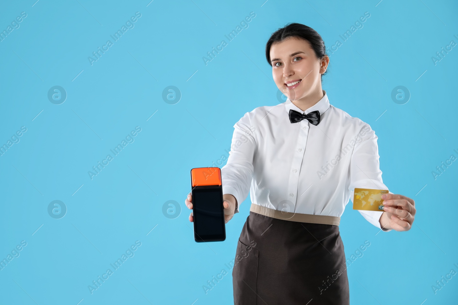 Photo of Happy waitress with payment terminal and credit card on light blue background, space for text