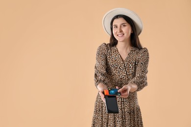 Happy young woman with payment terminal and debit card on beige background, space for text