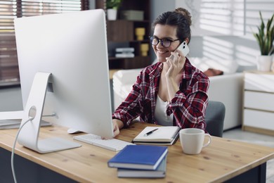 Photo of Happy woman talking by smartphone at desk with computer indoors. Home office