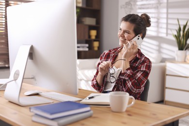 Photo of Happy woman talking by smartphone at desk with computer indoors. Home office