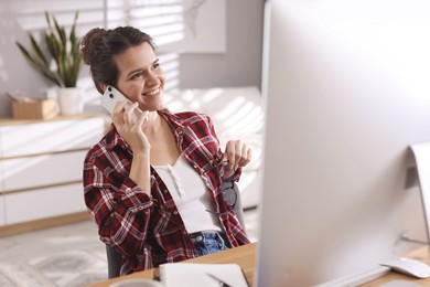 Photo of Happy woman talking by smartphone at desk with computer indoors. Home office