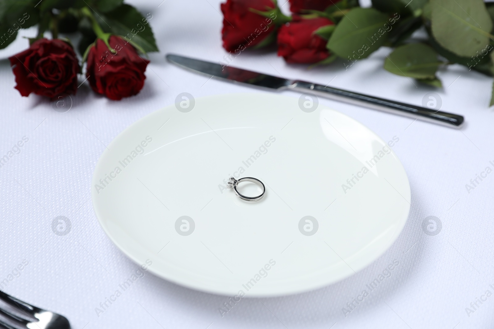 Photo of Engagement ring, rose flowers and tableware on white tablecloth, closeup. Table setting for romantic dinner