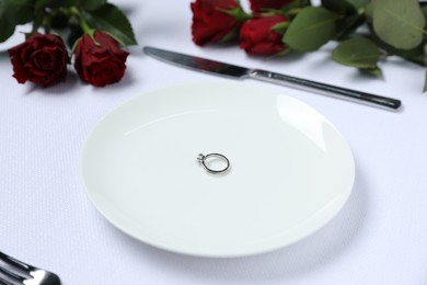 Engagement ring, rose flowers and tableware on white tablecloth, closeup. Table setting for romantic dinner