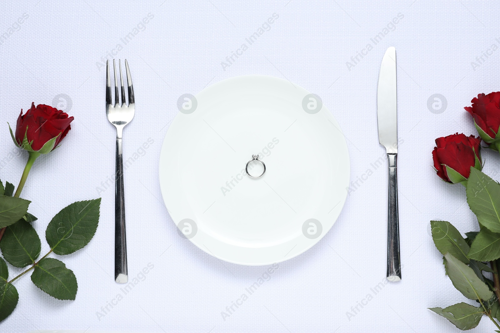 Photo of Engagement ring, rose flowers and tableware on white tablecloth, flat lay. Table setting for romantic dinner