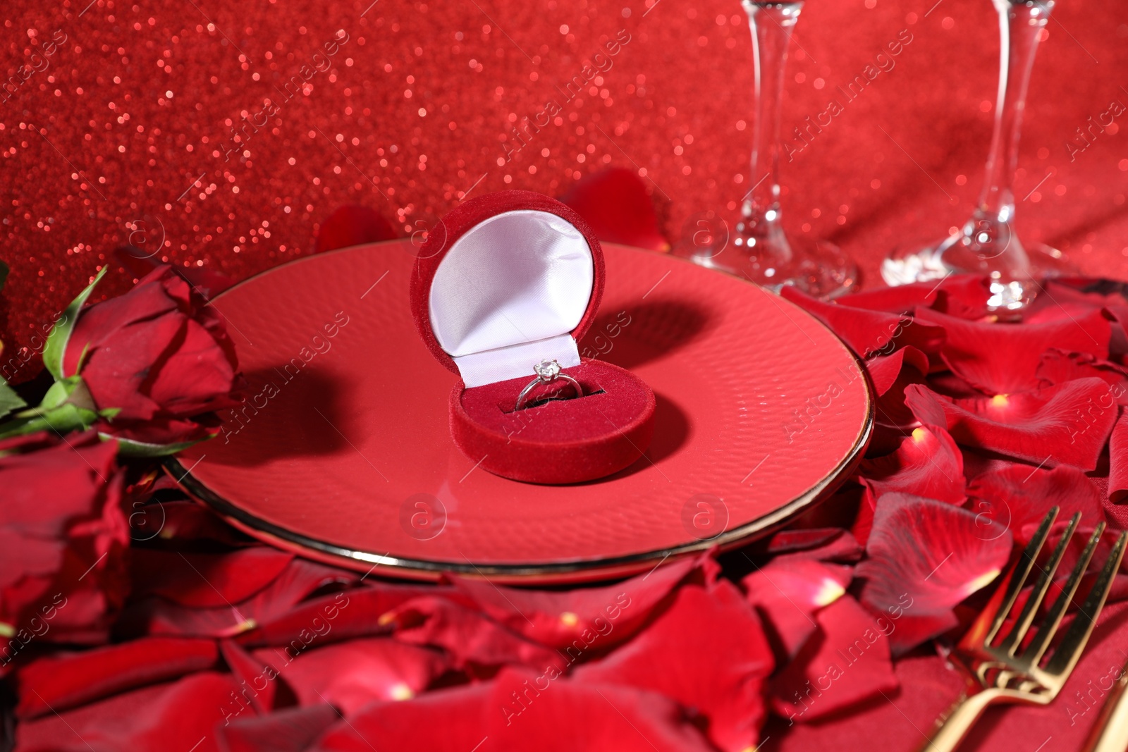 Photo of Engagement ring in box, tableware, rose flowers and petals on red glitter background, closeup. Table setting for romantic dinner