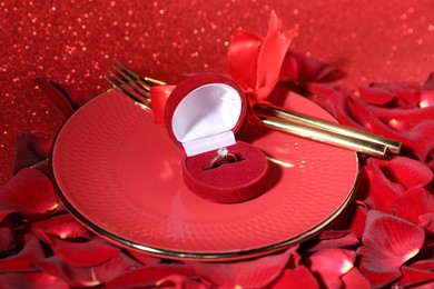 Photo of Engagement ring in box, tableware and rose petals on red glitter background, closeup. Table setting for romantic dinner