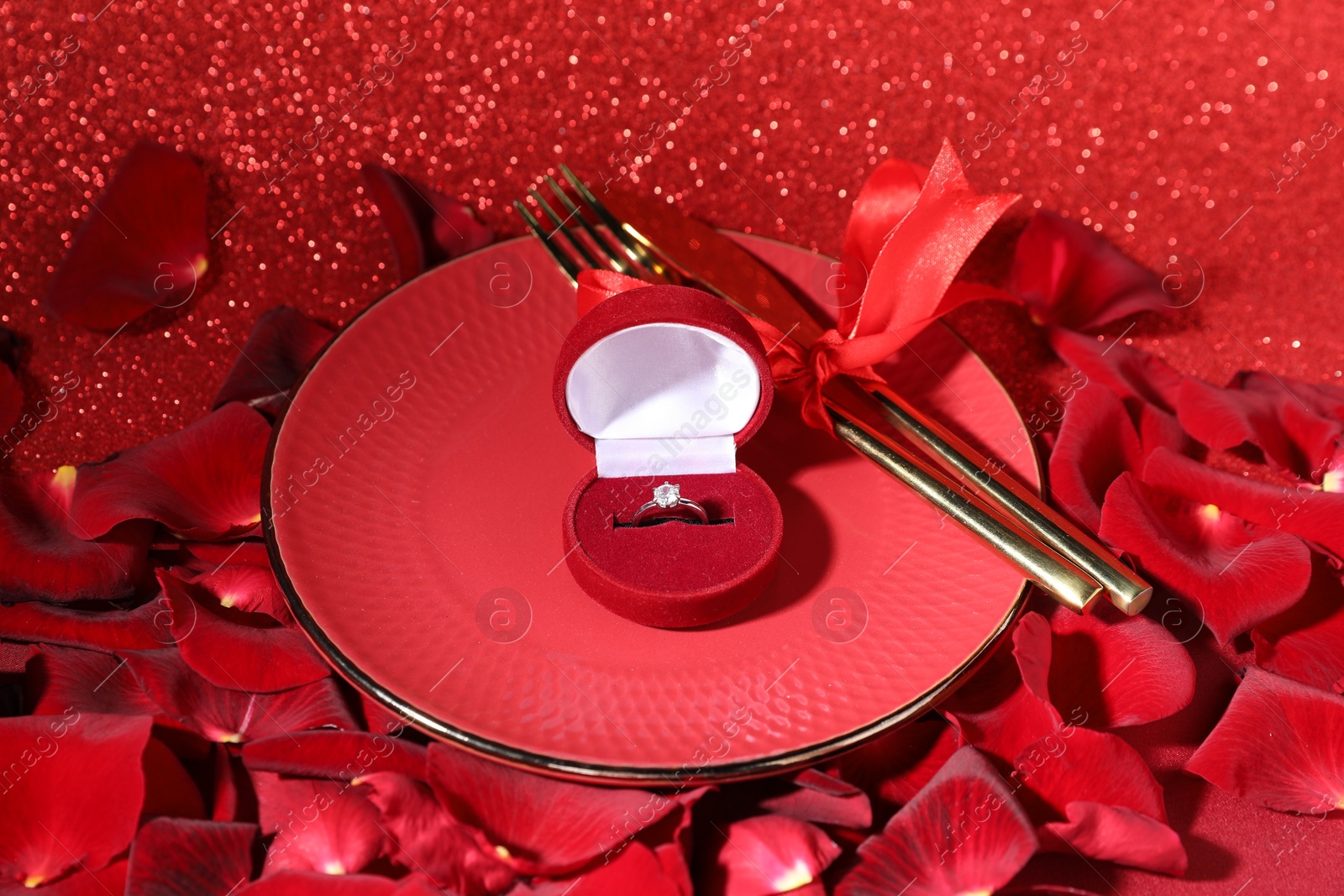 Photo of Engagement ring in box, tableware and rose petals on red glitter background, closeup. Table setting for romantic dinner