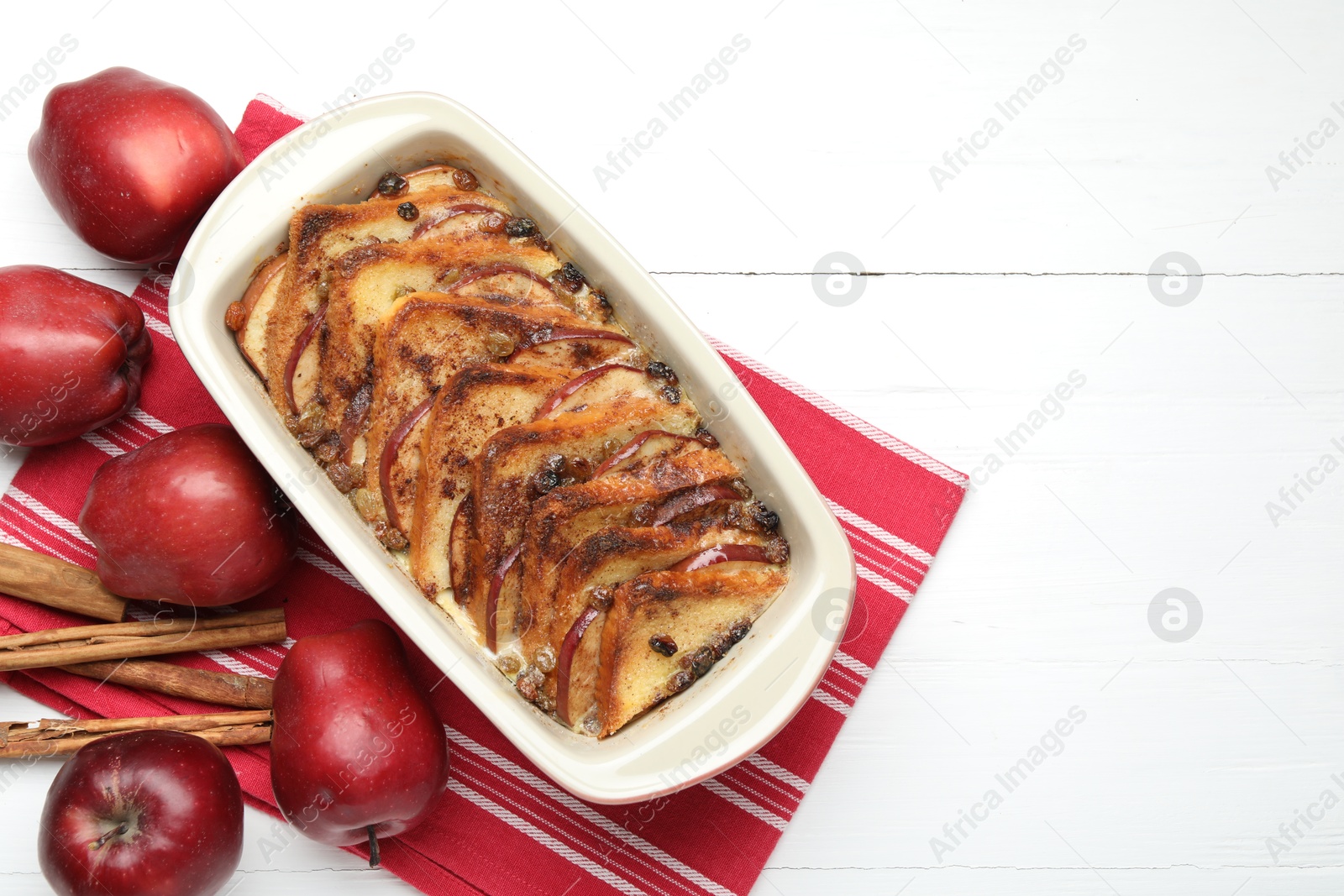 Photo of Delicious bread pudding in baking dish, fresh apples and cinnamon sticks on white wooden table, flat lay. Space for text