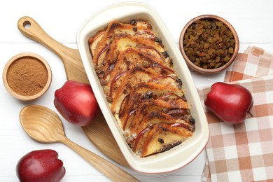 Photo of Delicious bread pudding in baking dish, fresh apples, cinnamon and raisins on white wooden table, flat lay