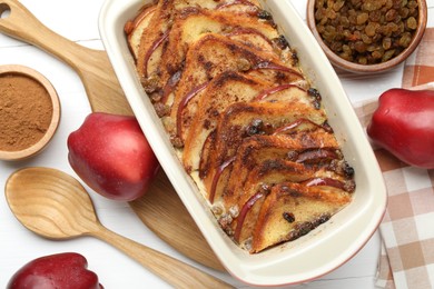 Photo of Delicious bread pudding in baking dish, fresh apples, cinnamon and raisins on white table, flat lay