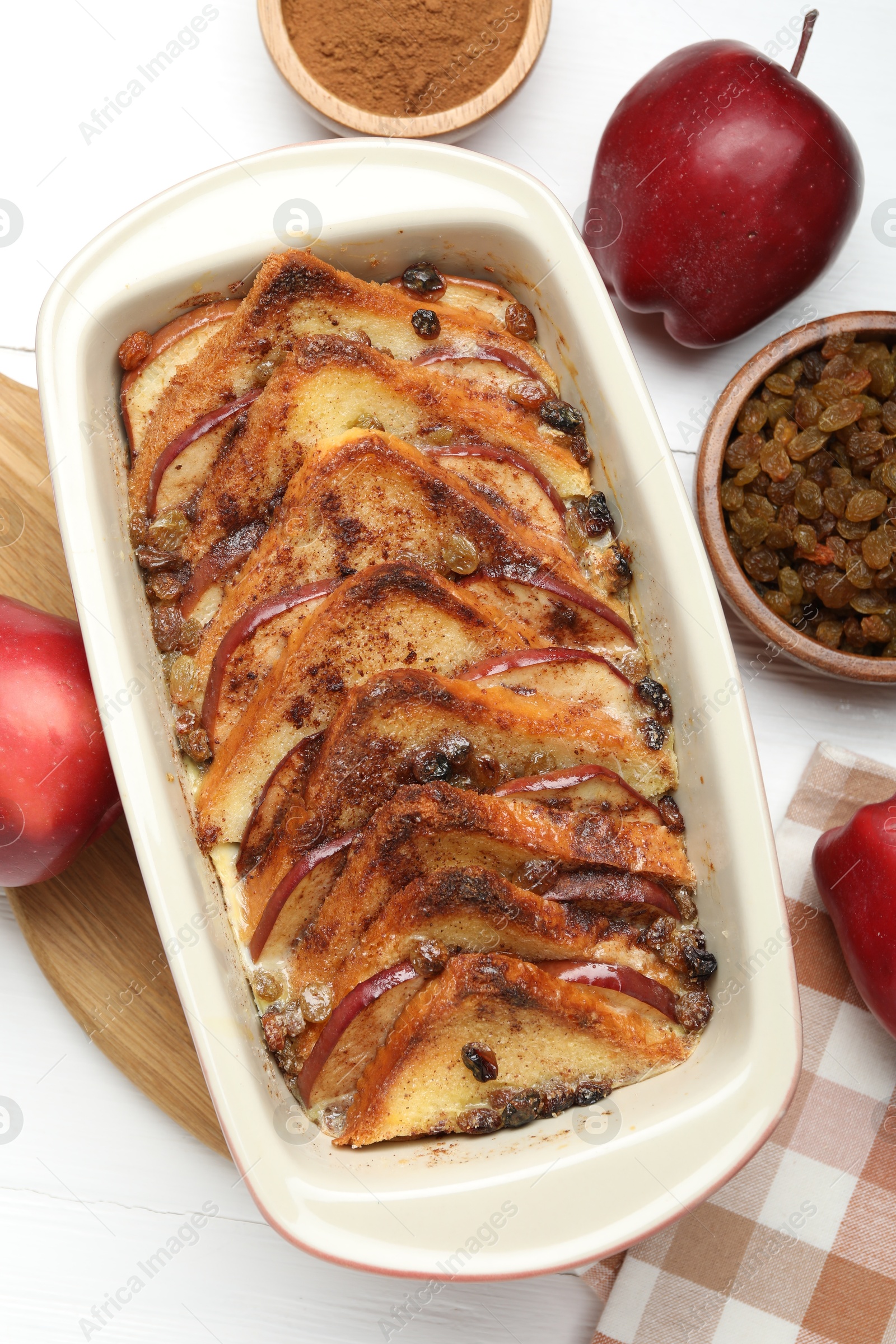 Photo of Delicious bread pudding in baking dish, fresh apples, cinnamon and raisins on white table, flat lay