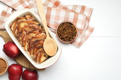 Photo of Delicious bread pudding in baking dish, fresh apples, cinnamon and raisins on white wooden table, flat lay. Space for text