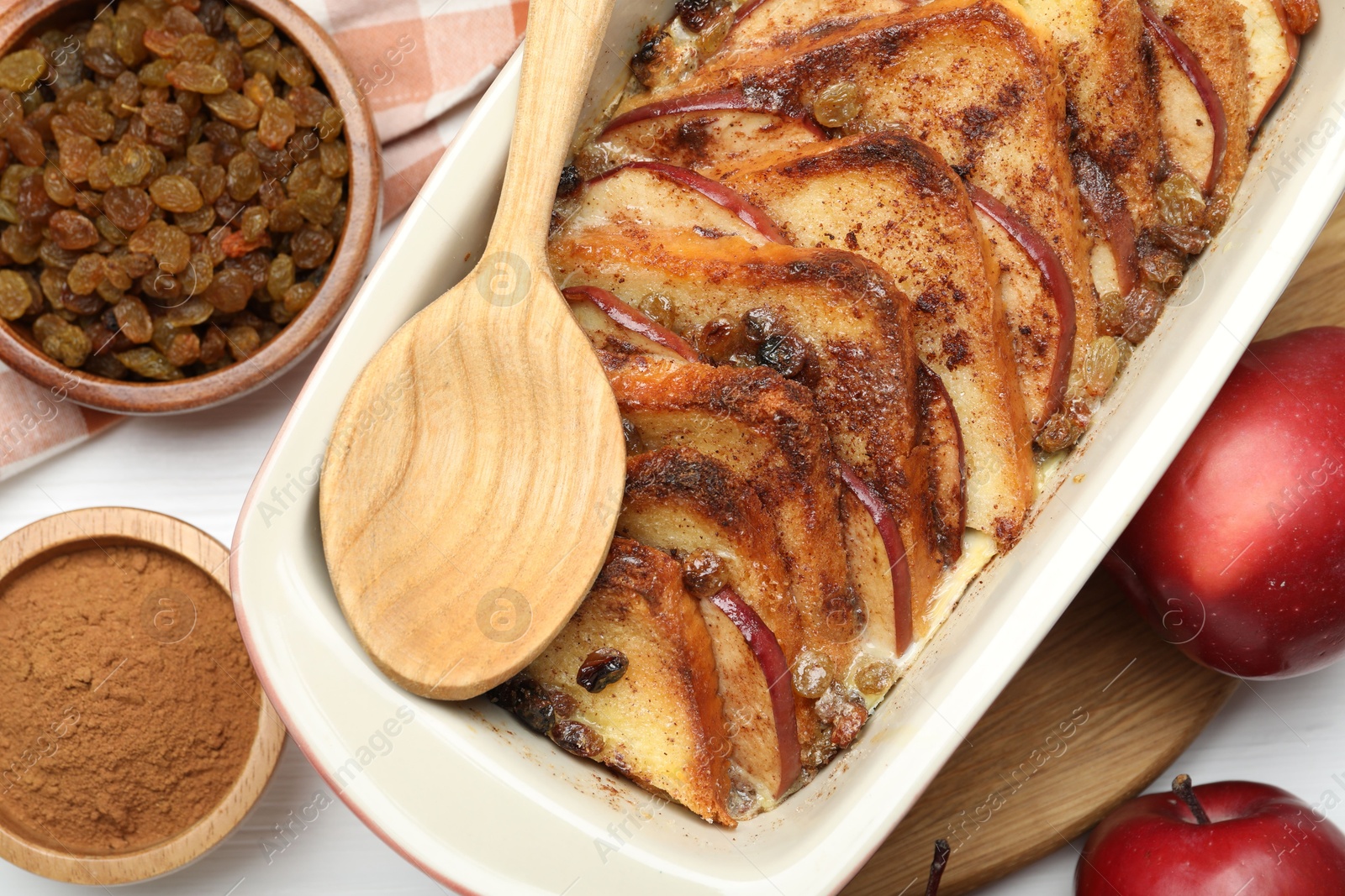 Photo of Delicious bread pudding in baking dish, fresh apples, cinnamon and raisins on white table, flat lay