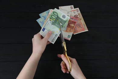 Photo of Woman cutting euro banknotes at black wooden table, top view