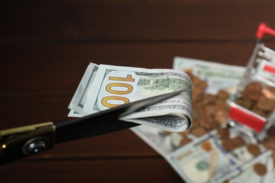 Photo of Scissors with dollar banknotes, coins and small shopping cart on brown table, selective focus