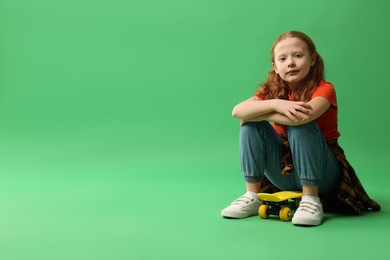 Photo of Stylish little girl sitting on penny board against green background, space for text