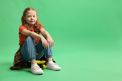 Photo of Stylish little girl sitting on penny board against green background, space for text