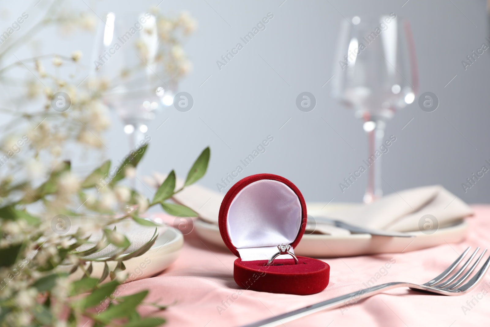 Photo of Engagement ring in box, branches with leaves and tableware on pink tablecloth, closeup. Romantic dinner