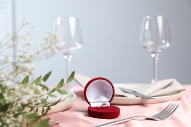 Photo of Engagement ring in box, branches with leaves and tableware on pink tablecloth, closeup. Romantic dinner