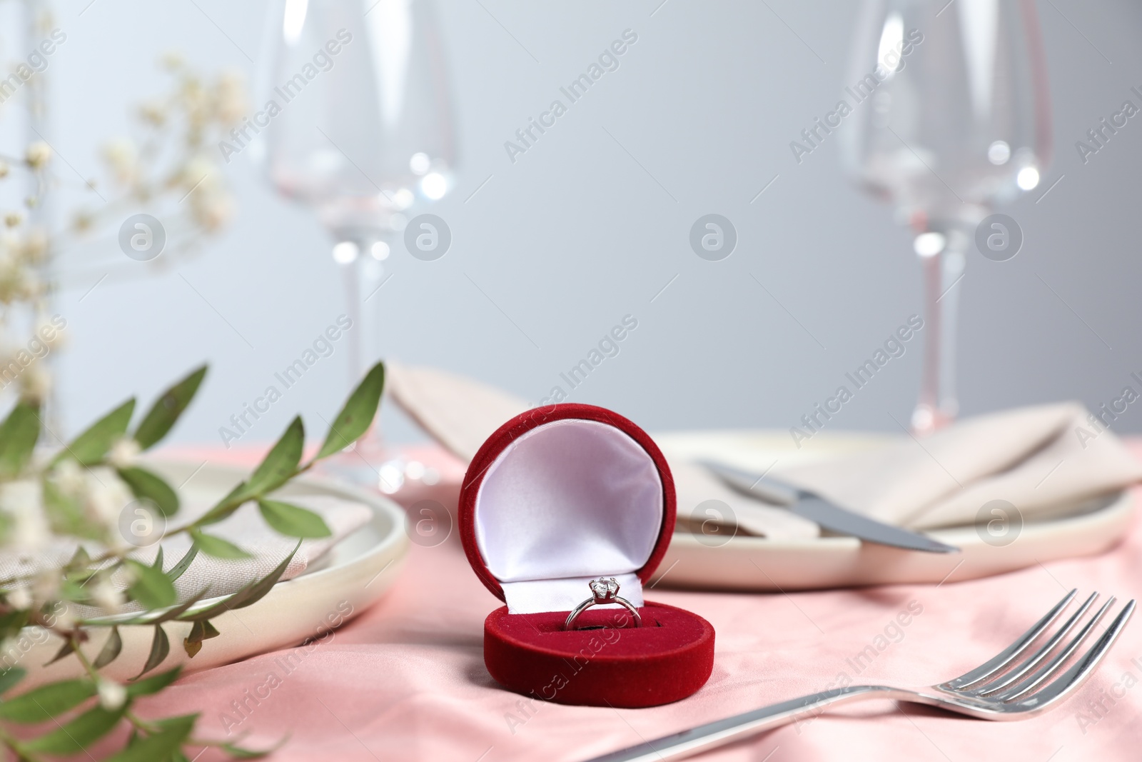 Photo of Engagement ring in box, branches with leaves and tableware on pink tablecloth, closeup. Romantic dinner