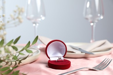 Photo of Engagement ring in box, branches with leaves and tableware on pink tablecloth, closeup. Romantic dinner