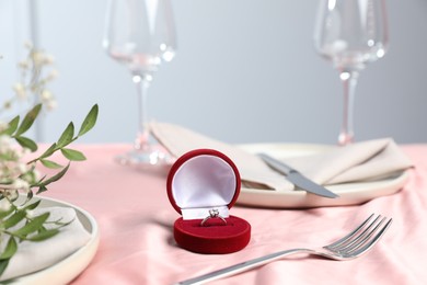 Photo of Engagement ring in box, branches with leaves and tableware on pink tablecloth, closeup. Romantic dinner