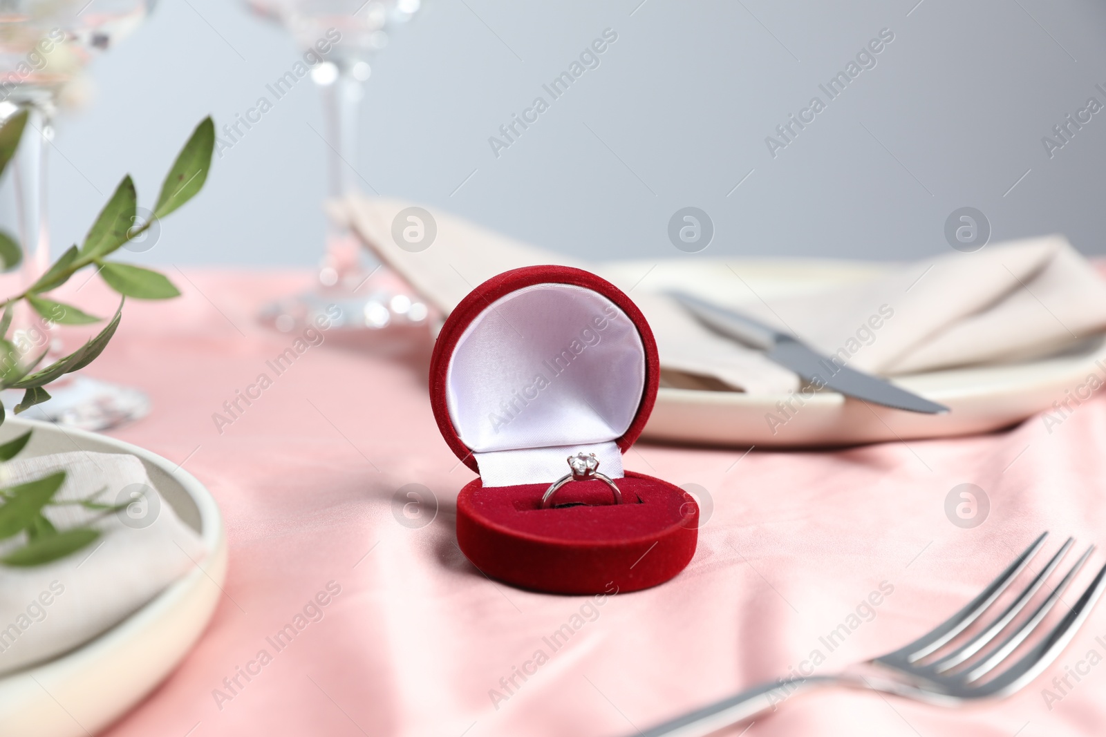 Photo of Engagement ring in box, branches with leaves and tableware on pink tablecloth, closeup. Romantic dinner