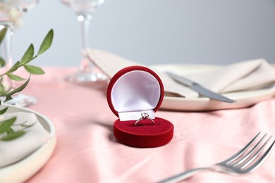 Engagement ring in box, branches with leaves and tableware on pink tablecloth, closeup. Romantic dinner