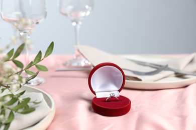 Photo of Engagement ring in box, branches with leaves and tableware on pink tablecloth, closeup. Romantic dinner