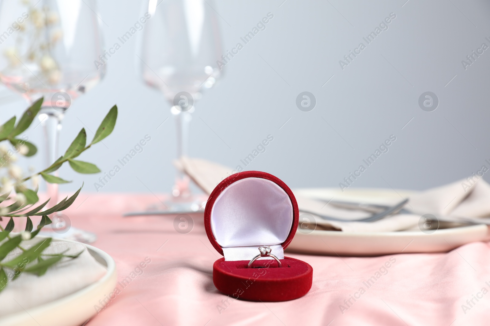 Photo of Engagement ring in box, branches with leaves and tableware on pink tablecloth, closeup. Romantic dinner