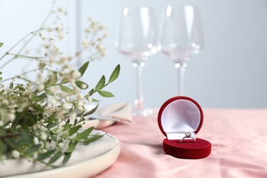 Photo of Engagement ring in box, branches with flowers and plate on pink tablecloth, closeup. Romantic dinner