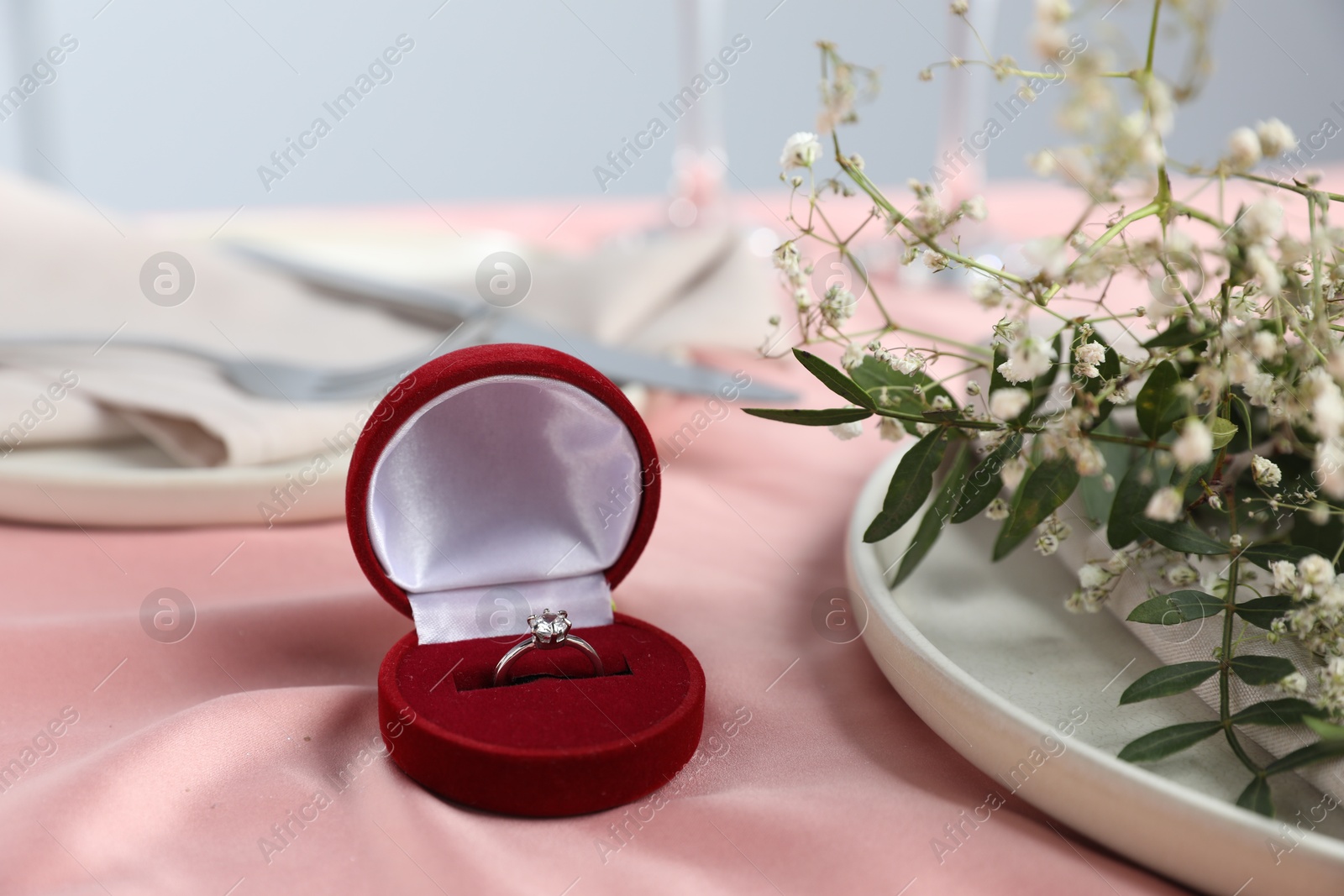 Photo of Engagement ring in box, branches with flowers and plate on pink tablecloth, closeup. Romantic dinner