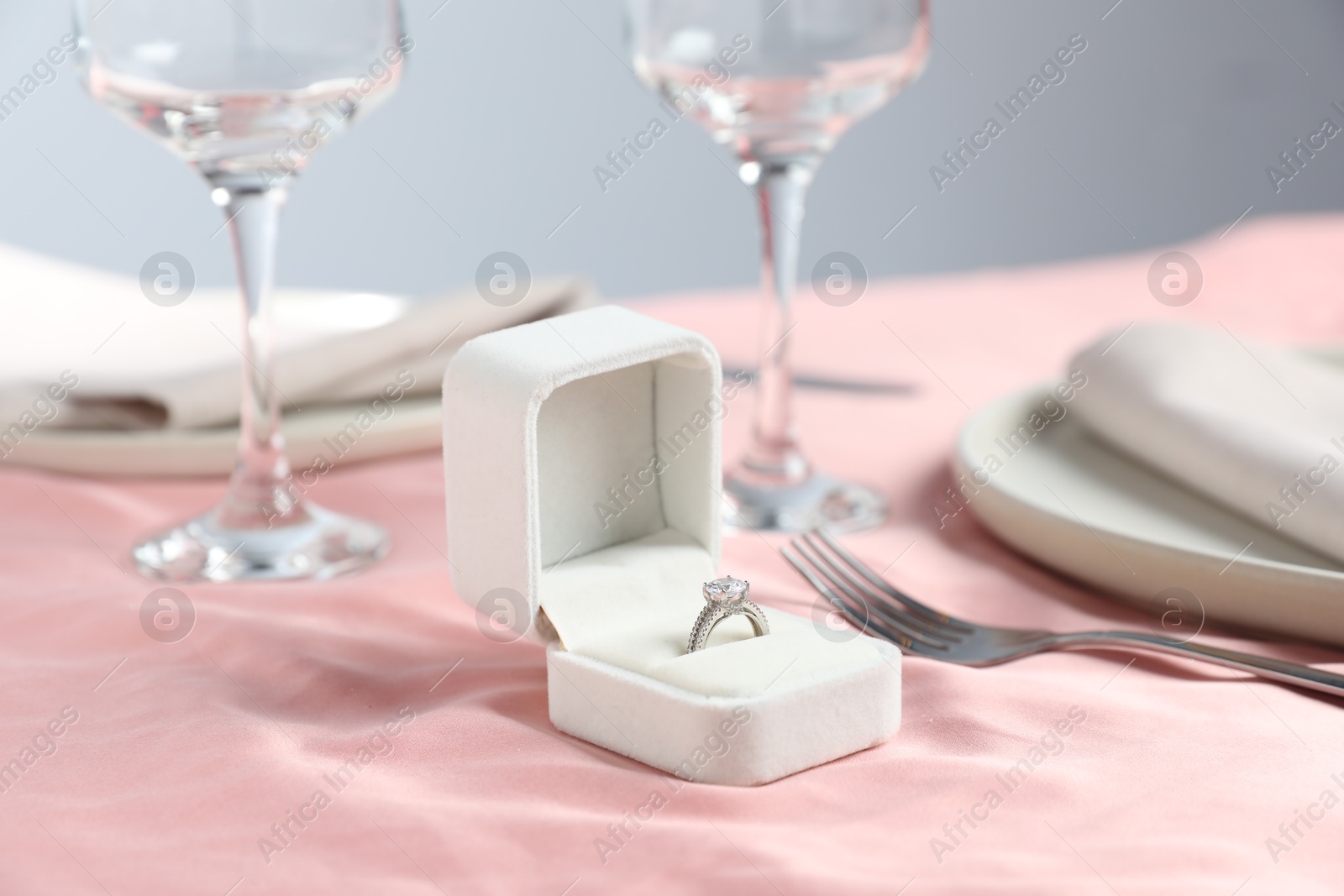 Photo of Engagement ring in box and tableware on pink tablecloth, closeup. Romantic dinner