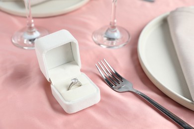 Engagement ring in box and tableware on pink tablecloth, closeup. Romantic dinner