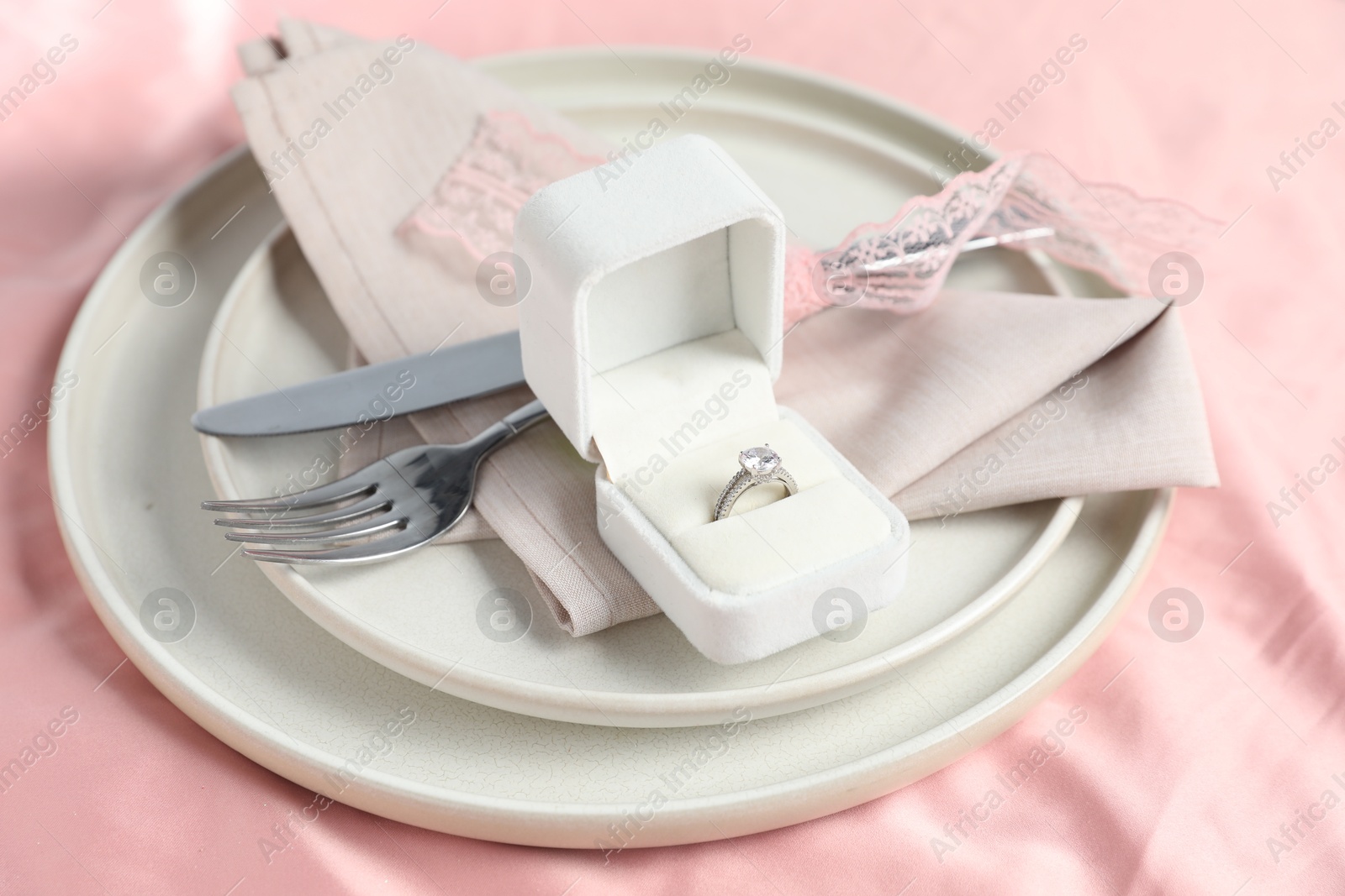 Photo of Engagement ring in box and tableware on pink tablecloth, closeup. Romantic dinner