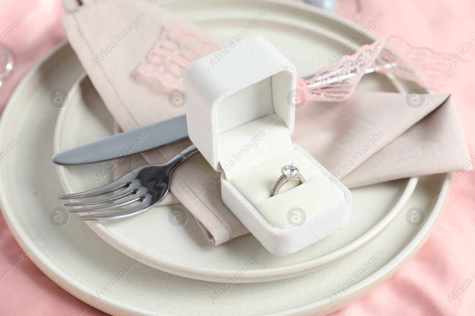 Photo of Engagement ring in box and tableware on pink tablecloth, closeup. Romantic dinner