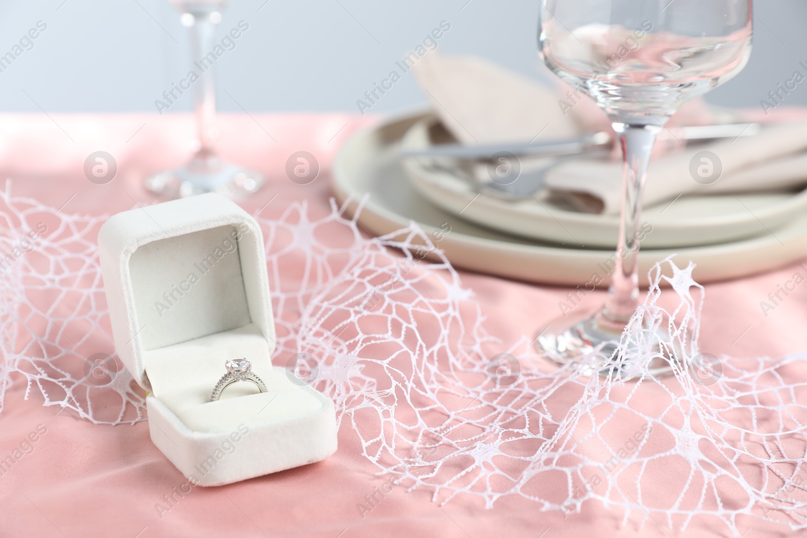 Photo of Engagement ring in box and tableware on pink tablecloth, closeup. Romantic dinner