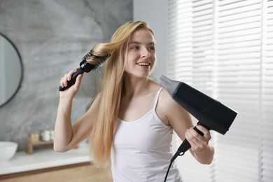 Photo of Beautiful young woman styling her hair with hairdryer and brush in bathroom