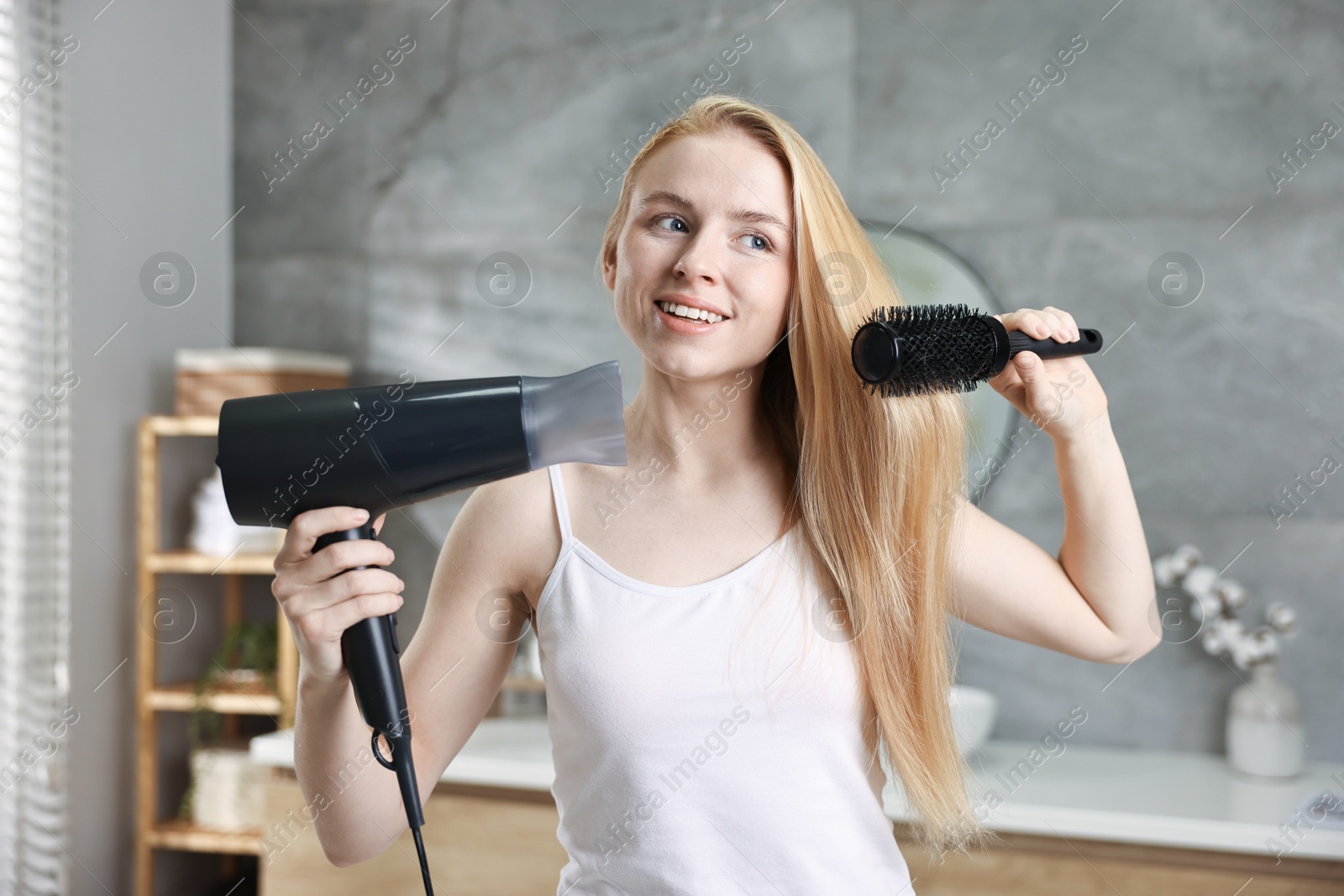 Photo of Beautiful young woman styling her hair with hairdryer and brush in bathroom