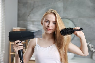 Photo of Beautiful young woman styling her hair with hairdryer and brush in bathroom