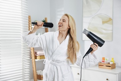 Photo of Beautiful young woman drying her hair and singing into brush in bathroom