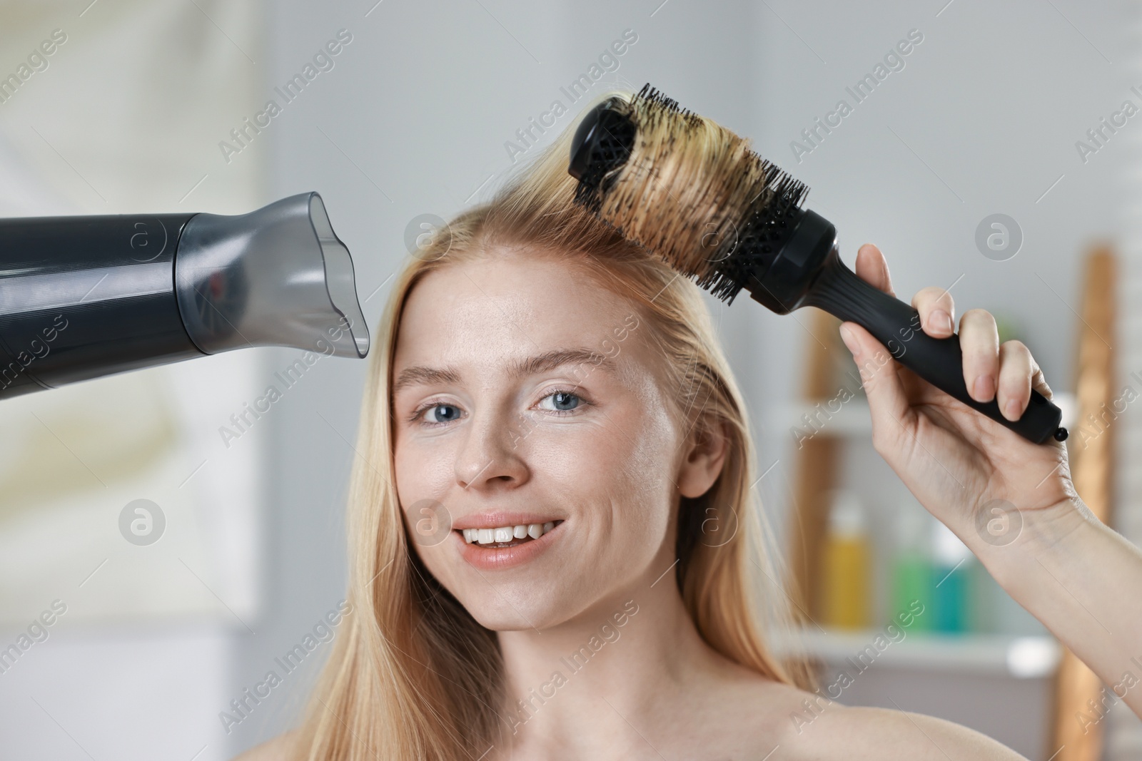 Photo of Beautiful young woman styling her hair with hairdryer and brush in bathroom