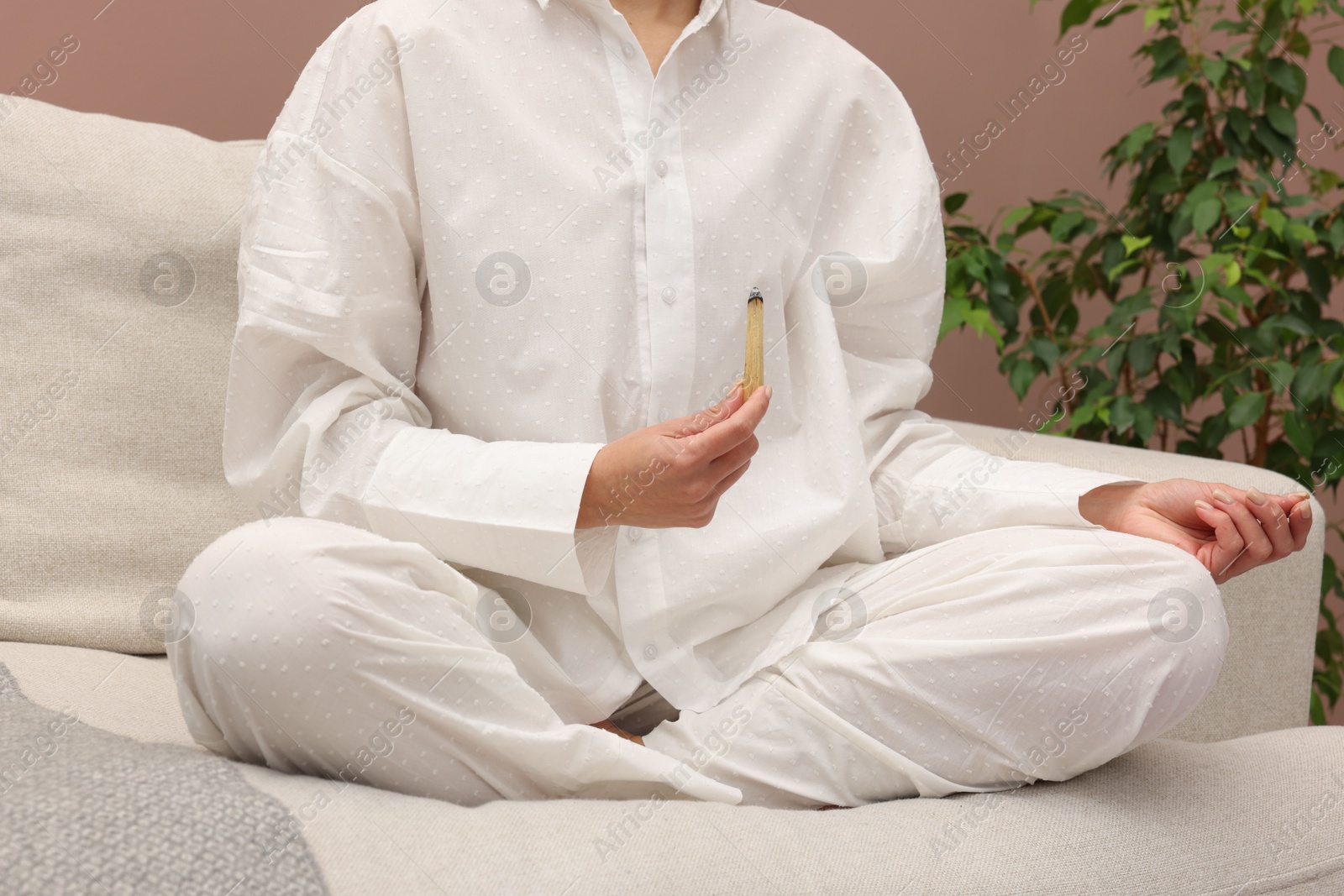 Photo of Woman with smoldering palo santo stick meditating on sofa at home, closeup