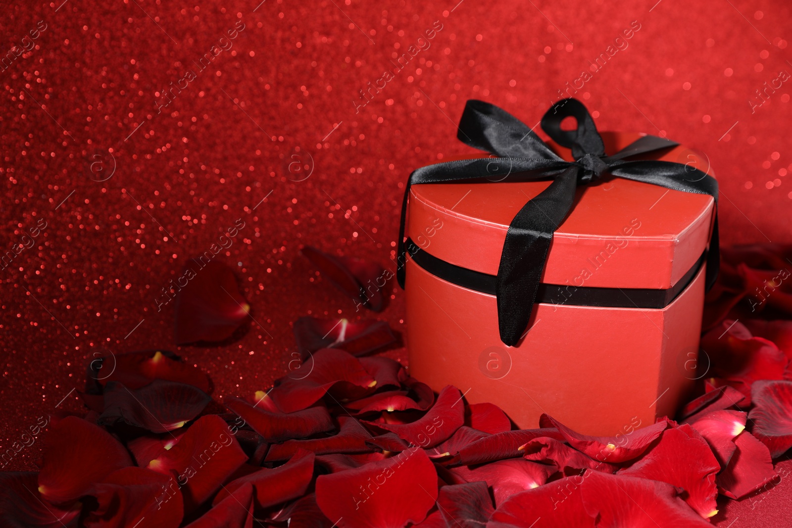 Photo of Beautiful gift box in shape of heart and rose petals on red glitter background, closeup with space for text. Happy Valentine's day